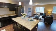 This two-floor residence offers a thoughtfully designed layout with modern finishes throughout. The kitchen serves as a central highlight, featuring dark espresso cabinetry, granite countertops, stainless steel appliances, and a large island with seating for two, illuminated by pendant lighting overhead. Rich hardwood-style flooring runs throughout the main level, complementing the kitchen's bold, contemporary palette.

The open-concept design connects the kitchen seamlessly to the living area, which benefits from multiple large windows that draw in generous natural light and provide views of the surrounding greenery. The dining space accommodates a tall pub-style table with industrial bar stools, adding a casual, functional element to the main floor.

Both bedrooms are carpeted and comfortably sized, each featuring large windows with tree-lined views, creating bright and restful retreats. A dedicated laundry room with a full-size washer and dryer adds everyday convenience to the home.

Neutral wall tones are consistent throughout, providing a clean, cohesive backdrop across all rooms. The overall ambiance is modern and welcoming, balancing practical functionality with tasteful design details.