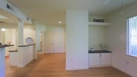 This single-floor space features a clean, open layout that promotes a sense of flow and functionality throughout. The interior is finished with warm-toned hardwood flooring that extends seamlessly across the main living areas, contributing to a cohesive and inviting ambiance. Neutral white walls and recessed ceiling lighting create a bright, airy atmosphere that feels both modern and welcoming.

A central structural column subtly defines distinct zones within the open floor plan without fully enclosing the space. On one side, a built-in shelving and cabinet unit with a dark countertop surface provides practical storage and display space, accented by a decorative "FAMILY" sign — adding a personal touch to the area. This built-in feature includes white cabinetry beneath, maintaining a consistent, clean aesthetic.

Adjacent to the main living area, a glimpse of what appears to be a kitchen is visible, featuring dark countertops that complement the overall design palette. A hallway leads toward additional rooms, with a door visible in the background, suggesting further private spaces beyond the main living zone. Overall, the space presents a practical and neatly finished interior suited for everyday living.