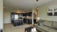 This single-floor space showcases an open-concept layout that seamlessly connects the kitchen and living area, creating a cohesive and functional environment. The kitchen is a standout feature, equipped with dark espresso cabinetry that extends to the ceiling, providing ample storage. Stainless steel appliances, including a refrigerator, stove, microwave, and dishwasher, complement the cabinetry. A kitchen island with seating for two offers additional prep space and casual dining. Track lighting overhead ensures the kitchen remains well-lit and practical.

The living area flows naturally from the kitchen, anchored by neutral-toned carpeting that adds warmth underfoot. A ceiling fan overhead provides comfort and air circulation. The living space is furnished with a plush sofa accented with decorative pillows and a dark coffee table, contributing to a relaxed yet polished atmosphere. Neutral wall tones throughout unify the space and enhance its open, airy feel. A glimpse into an adjacent hallway suggests additional rooms beyond, adding to the overall functionality of this well-designed layout.