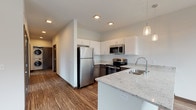 This single-floor unit showcases a contemporary design with a cohesive palette of white walls, dark wood-toned accents, and durable wood-look flooring throughout the main living areas. The open-concept kitchen serves as a central feature, highlighted by a generous island with light granite countertops, an undermount double sink, and elegant pendant lighting overhead. Cabinetry combines upper white units with darker lower cabinets, complemented by a full suite of stainless steel appliances including a refrigerator, range, microwave, and dishwasher. A large window beside the sink and additional floor-to-ceiling glazing flood the kitchen with natural light and outdoor views.

The living area extends from the kitchen and offers sliding glass door access to an outdoor space, with additional windows enhancing the bright, airy atmosphere. Bedroom spaces feature plush carpeting, ceiling fans, windows providing natural light, and closet storage. Two bathrooms are finished with dark-toned vanity cabinetry, white quartz countertops, and clean tile flooring — one with a walk-in shower, the other with a tub-shower combination. A dedicated hallway houses a stacked in-unit washer and dryer, adding everyday convenience to this well-appointed space.