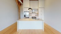 Located at 326 W Florida St in Milwaukee's vibrant Walker's Point neighborhood, this single-floor loft masterfully blends historic industrial character with contemporary design. The open-concept layout is anchored by striking exposed wood beam ceilings and original timber post-and-beam framing, which contrast beautifully against crisp white walls and smooth light hardwood flooring that runs throughout the space.

The kitchen serves as a focal point, featuring a large waterfall-edge island with an integrated sink, sleek white cabinetry extending to ceiling height, stainless steel appliances including a French door refrigerator and range, a wall-mounted range hood, and a classic subway tile backsplash with open wood shelving accents. A bold pendant light anchors the island, adding a modern industrial touch.

The main living area is generously proportioned, offering an expansive, flexible layout bathed in natural light from tall arched windows framed by exposed brick — a nod to the building's historic roots. Views of the surrounding Milwaukee neighborhood are visible through these architecturally distinctive windows.

The bathroom is clean and functional, finished with light gray tile flooring, accessible grab bars, a soaking tub, and a glass-enclosed shower, maintaining the space's refined, modern aesthetic. The overall ambiance is bright, airy, and full of character.