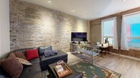 Located at 123 King Street in downtown London, Ontario, this one-bedroom, one-bathroom unit offers a thoughtfully designed layout that maximizes its compact footprint. The open-concept kitchen and living area serves as the heart of the space, where a well-appointed kitchen stands out with crisp white shaker-style cabinetry, marble-toned countertops, stainless steel appliances including a range and refrigerator, and eye-catching under-cabinet purple accent lighting. A full-height bookshelf adds character and function near the kitchen. The living area is anchored by a striking full-height exposed brick feature wall that lends an urban, industrial warmth to the space, complemented by light hardwood flooring and recessed ceiling lighting throughout. A sectional leather sofa, area rug, and media setup make this area both comfortable and inviting, with a window allowing natural light to filter in. The bedroom continues the exposed brick aesthetic and features double doors with a transom window above, adding architectural interest. The bathroom is clean and neutral, finished with tile flooring, a wall-mounted storage cabinet, and simple, functional fixtures. A connecting hallway ties the rooms together efficiently. This unit blends modern updates with timeless character details for a distinctive urban living experience.