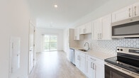 This single-floor unit is designed with a clean, modern aesthetic throughout, characterized by crisp white walls, light wood-tone flooring, and an abundance of natural light. The open-concept layout connects the kitchen directly to the living area, creating a seamless flow that enhances the sense of space.

The kitchen is well-appointed with white shaker-style cabinetry, light quartz countertops, and a distinctive hexagonal tile backsplash. Stainless steel appliances — including a range, over-the-range microwave, dishwasher, and refrigerator — provide full functionality. The living area extends toward a sliding glass door that opens to a private balcony overlooking lush greenery, filling the interior with natural light.

The bedroom is generously sized with vaulted ceiling detail near the entrance and access to a walk-in closet fitted with wire shelving for organized storage. A second door leads to a sunlit secondary space or additional room.

The bathroom features a white shaker vanity with quartz countertops, an undermount sink, chrome hardware, and a framed mirror, maintaining the cohesive, contemporary palette found throughout the unit. Light wood flooring runs consistently across all rooms, reinforcing the bright and unified design.