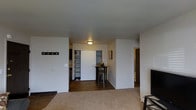 This single-floor space is designed with an open-concept layout that maximizes functionality and flow. The living area features plush wall-to-wall carpeting, creating a warm and comfortable atmosphere, while the transition into the kitchen and dining zone is marked by hardwood-style flooring that adds a clean, practical finish.

The kitchen area is efficiently arranged and connects seamlessly to a compact dining nook, which features a bistro-style high table with bar stools — an ideal spot for casual meals or a workspace. A built-in storage closet with open shelving provides generous organizational capacity, keeping the space tidy and well-utilized.

A coat and accessory rack mounted near the entry area offers a convenient drop zone upon arrival, enhancing the practical design of the unit. The neutral color palette — soft whites and warm beiges throughout the walls and ceilings — contributes to a bright and airy ambiance despite the compact footprint.

Flush-mount ceiling lighting provides consistent illumination across the main living and kitchen areas. A hallway visible in the background suggests access to additional private rooms, completing this thoughtfully arranged single-floor layout that balances comfort and practicality.