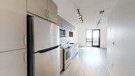 This single-floor unit presents a contemporary design characterized by clean lines, light hardwood flooring, and a bright, open layout throughout. The kitchen is thoughtfully appointed with flat-front cabinetry in a warm taupe finish, a white subway tile backsplash, and a full suite of stainless steel appliances, including a refrigerator, electric range, built-in microwave, and dishwasher. A sleek white countertop with an undermount sink completes the functional workspace, illuminated by adjustable track lighting overhead.

The open living area flows seamlessly from the kitchen, offering generous space bathed in natural light. A set of dark-framed glass doors leads to a private balcony, providing outdoor views and an airy ambiance. A ceiling fan is installed overhead, adding to the comfort of the space.

A well-sized closet with double hanging rods provides ample built-in storage. The bathroom features a tan vanity with a white quartz countertop, a large mirror with contemporary bar lighting, built-in shelving, and a tub-shower combination. Throughout, white walls and light flooring create a cohesive, modern aesthetic.