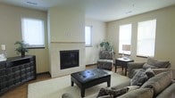 This single-floor space offers a thoughtfully designed living area that combines comfort and modern style. The focal point of the room is a tile-surround fireplace, framed by clean, neutral-toned walls that create a warm and inviting atmosphere. The fireplace's light-colored tile surround extends to the ceiling, adding a sense of architectural height and structure to the space.

Natural light fills the room through multiple windows fitted with horizontal blinds, providing both brightness and privacy. The flooring features warm hardwood tones, partially covered by a plush, cream-colored area rug that anchors the seating arrangement. The living area is furnished with a coordinated set of upholstered sofas and accent chairs in muted, earthy tones, complemented by a tufted leather ottoman that serves as a central coffee table.

A dedicated seating nook adjacent to the main living area offers a cozy retreat, complete with a patterned accent chair, a dark wood side table, and a table lamp. A decorative potted plant adds a touch of greenery, enhancing the space's welcoming ambiance. The overall design balances functionality with aesthetic appeal, making it a comfortable and well-appointed living environment.