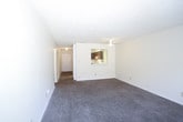 This single-floor unit offers a straightforward, functional layout across several well-defined spaces. The living area is generously sized, finished with wall-to-wall brown carpet and neutral cream-toned walls. A ceiling fan with an integrated light fixture provides both ventilation and ambient lighting. A sliding glass door fitted with vertical blinds opens to an outdoor area, allowing natural light to filter into the room and providing a connection to the exterior. The textured popcorn ceiling is consistent throughout the carpeted areas of the unit.

The bedroom features the same carpet and neutral walls, with a standard window that brings in natural light and exterior views of surrounding greenery. A built-in closet with rod and shelf storage is accessible via bifold doors, and an additional doorway connects to the rest of the unit.

The kitchen is equipped with dark wood cabinetry, dark countertops, a white electric range with an overhead ventilation hood, a dishwasher, and a tiled backsplash. The floor transitions to stone-look tile, which also carries into the bathroom. The bathroom includes a full tub-shower combination with a white surround, a toilet, and a vanity, all finished in a clean, neutral palette.
