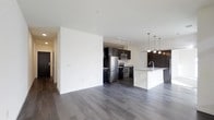 This single-floor space features an open-concept layout that seamlessly connects the living area to the kitchen, creating a sense of spaciousness and flow. The kitchen is well-appointed with dark espresso cabinetry that provides a striking contrast against the white walls and light-toned granite or stone countertops. Stainless steel appliances, including a refrigerator, stove, and built-in microwave, lend a modern, polished aesthetic. A central island with an integrated sink serves as both a functional workspace and a visual anchor for the kitchen, illuminated by a row of pendant lights hanging overhead. Track lighting above the main kitchen area ensures ample task lighting. The open living area is characterized by clean white walls and smooth ceilings, offering a neutral canvas adaptable to various design styles. Rich, dark hardwood-style flooring runs throughout the entire space, grounding the interior with warmth and continuity. A hallway visible on the left suggests additional rooms beyond the main living area. Overall, the space presents a contemporary, well-designed interior with a cohesive color palette of dark and light tones.