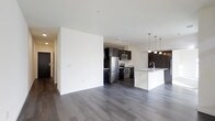This single-floor space features an open-concept layout that seamlessly connects the living area to the kitchen, creating a sense of spaciousness and flow. The kitchen is well-appointed with dark espresso cabinetry that provides a striking contrast against the white walls and light-toned granite or stone countertops. Stainless steel appliances, including a refrigerator, stove, and built-in microwave, lend a modern, polished aesthetic. A central island with an integrated sink serves as both a functional workspace and a visual anchor for the kitchen, illuminated by a row of pendant lights hanging overhead. Track lighting above the main kitchen area ensures ample task lighting. The open living area is characterized by clean white walls and smooth ceilings, offering a neutral canvas adaptable to various design styles. Rich, dark hardwood-style flooring runs throughout the entire space, grounding the interior with warmth and continuity. A hallway visible on the left suggests additional rooms beyond the main living area. Overall, the space presents a contemporary, well-designed interior with a cohesive color palette of dark and light tones.