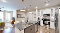 This well-appointed one-bedroom, one-bathroom unit in Grapevine, Texas offers a thoughtfully designed layout that maximizes its comfortable footprint. The open-concept kitchen and living area serves as the heart of the space, where the kitchen stands out with white shaker cabinetry, glass-front upper shelves, a subway tile backsplash, and a full suite of stainless steel appliances. A large center island with granite countertops, a deep undermount sink, and a matte black faucet provides both prep space and seating for multiple bar stools. Gold pendant lights overhead add a refined, contemporary touch. A sliding barn door connects the main living area to the hallway, lending a stylish architectural detail. The living room is bright and welcoming, featuring wood-look flooring, a ceiling fan, and French doors that open to an outdoor area. The bedroom is a calm retreat with natural light from dual windows, a ceiling fan, and direct access to the bathroom. The bathroom includes a combined tub and shower with neutral tile surroundings and wood-look flooring. A dedicated walk-in closet with built-in shelving and hanging rods completes the private quarters, offering practical and organized storage throughout.