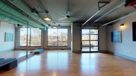 Located at 1300 N Fordham Blvd in Chapel Hill, NC, this single-floor fitness and studio space combines an industrial-modern aesthetic with a bright, functional layout. The main studio area is expansive and open, featuring warm cork flooring that provides both durability and comfort underfoot. Striking teal-painted exposed ductwork runs across the ceiling, lending a distinctive industrial character to the space, while pendant and track lighting ensure the room is well-lit throughout the day.

Large, floor-to-ceiling windows line the walls, flooding the interior with abundant natural light and offering unobstructed views of the surrounding landscape and neighboring buildings. A full-length glass panel garage door adds versatility and visual openness, connecting the studio to the outdoors. Wall-mounted mirrors enhance the sense of depth and are well-suited for movement-based activities.

A glass-partitioned entryway leads from the studio into a reception or lobby area, finished with tile flooring and branded signage, creating a clean separation between the public-facing front and the main activity floor. Additional private rooms or changing areas are accessible through light wood panel doors along one wall. The overall ambiance is airy, modern, and purposefully designed to support fitness, wellness, or creative studio programming.