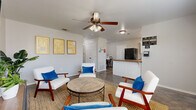 This well-appointed single-story space offers a thoughtfully organized layout designed for both comfort and functionality. The main lounge area serves as the heart of the space, featuring tile flooring, a ceiling fan with light fixture, and large windows that invite generous natural light. Mid-century modern seating with white upholstery and bold blue accent pillows is arranged around a rustic round coffee table, creating an inviting and relaxed atmosphere. A wall-mounted television and console table complete the living area.

The kitchen is efficiently designed with white cabinetry, warm countertops, stainless steel appliances including a full-size refrigerator, range, and dishwasher, and a window overlooking the exterior. An adjacent reception counter connects the kitchen area to the lounge.

A private office features a large wood L-shaped desk, filing cabinet, and a window with outdoor views, making it a productive and comfortable workspace. The bedroom is quietly appointed with neutral tones, carpeted floors, natural light from a window, and double closet doors offering ample storage.

Two bathrooms are finished with cream cabinetry, dark hardware, warm-toned countertops, framed mirrors, and polished concrete-style flooring, lending a cohesive and understated aesthetic throughout the space.