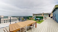 Perched above downtown Memphis, this rooftop lounge delivers an expansive open-air experience defined by sweeping views of the Mississippi River, the iconic Hernando de Soto Bridge, and the surrounding city skyline. The deck is finished in grey composite wood planking and organized into several distinct seating zones, each with its own character and purpose.

A generously sized lounge area anchors one section of the deck, featuring wicker sectional sofas with bold navy blue cushions arranged atop a blue-and-white geometric-patterned area rug, complemented by contemporary white pedestal side tables. Adjacent to this, patches of vibrant artificial turf introduce a lively green contrast to the neutral deck, hosting a teak dining table with white and wicker chairs — ideal for al fresco dining. Additional intimate seating vignettes with teal accent chairs and small round tables are scattered throughout, providing flexible social settings.

Green hedge privacy panels serve as both decorative dividers and windbreaks, creating a sense of enclosure without sacrificing the open-air atmosphere. A structure along one edge features green-and-white striped awnings and large windows, adding architectural charm. The railing perimeter throughout the deck ensures unobstructed sightlines across the Memphis skyline. This rooftop lounge is a well-appointed urban retreat suited for gatherings, relaxation, and taking in the distinctive character of the city below.