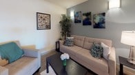This thoughtfully designed model unit offers a compelling look at contemporary urban living across a single floor. The living area greets visitors with polished concrete flooring, a natural wood-accented entry door, and a comfortable seating arrangement complemented by bold abstract artwork and an accent wall in a cool gray tone, creating a stylish yet inviting atmosphere.

The kitchen is a standout feature, outfitted with warm maple cabinetry, dark stone countertops, and a full suite of stainless steel appliances including a refrigerator, range with microwave overhead, and dishwasher. Track lighting above and a pendant light over the sink area add a refined, functional touch, while the generous counter and cabinet space makes the kitchen as practical as it is attractive.

The bedroom is a serene retreat, featuring large floor-to-ceiling windows with vertical blinds that invite abundant natural light and offer expansive outdoor views. A convenient in-unit stacked washer and dryer is tucked neatly within the room, adding everyday practicality. The bathroom is clean and modern, featuring maple cabinetry with a dark countertop, polished concrete flooring, and a shower-tub combination. Together, these spaces present a cohesive, well-considered design ideal for modern living.