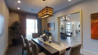 This single-floor space showcases a thoughtfully designed interior with a strong emphasis on elegant dining and entertaining. The primary dining area is anchored by a striking live-edge wood dining table, surrounded by plush upholstered chairs that invite leisurely gatherings. Above the table hangs a bold, large-scale rectangular pendant light with a woven rattan shade, casting a warm, textured glow that serves as a dramatic focal point. Decorative candle holders and a table runner add layers of sophistication to the tablescape. Rich, patterned drape curtains frame the windows, complementing the neutral, warm-toned walls and contributing to the space's refined character. A piece of contemporary artwork adorns the wall, adding a pop of color and artistic flair. An open archway connects the main dining area to a secondary dining or sitting room, which features its own chandelier, additional seating, and large windows that invite natural light. The continuous flow between the two spaces creates a sense of openness and versatility, making the layout well-suited for both intimate dinners and larger social gatherings. Overall, the space exudes a warm, upscale ambiance with a seamless blend of natural materials, layered lighting, and curated décor.