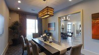This single-floor space showcases a thoughtfully designed interior with a strong emphasis on elegant dining and entertaining. The primary dining area is anchored by a striking live-edge wood dining table, surrounded by plush upholstered chairs that invite leisurely gatherings. Above the table hangs a bold, large-scale rectangular pendant light with a woven rattan shade, casting a warm, textured glow that serves as a dramatic focal point. Decorative candle holders and a table runner add layers of sophistication to the tablescape. Rich, patterned drape curtains frame the windows, complementing the neutral, warm-toned walls and contributing to the space's refined character. A piece of contemporary artwork adorns the wall, adding a pop of color and artistic flair. An open archway connects the main dining area to a secondary dining or sitting room, which features its own chandelier, additional seating, and large windows that invite natural light. The continuous flow between the two spaces creates a sense of openness and versatility, making the layout well-suited for both intimate dinners and larger social gatherings. Overall, the space exudes a warm, upscale ambiance with a seamless blend of natural materials, layered lighting, and curated décor.