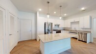 This thoughtfully designed single-floor unit offers a clean, modern aesthetic throughout, with warm wood-look flooring and crisp white walls creating a bright, welcoming atmosphere. The kitchen serves as a centerpiece of the space, showcasing white shaker cabinetry, granite countertops, and a full suite of stainless steel appliances including a French door refrigerator, electric range, and dishwasher. A central island with a granite surface provides additional prep space and is highlighted by stylish pendant lighting overhead. The flooring in the kitchen and living areas features a striking herringbone pattern that adds visual interest and elegance. The living and dining areas are spacious and open, benefiting from natural light and a seamless flow between rooms. The primary bedroom is generously sized, with a tray ceiling, ceiling fan, and windows that bring in outdoor views and natural light. The en-suite bathroom is well-appointed, featuring a dual-sink vanity with granite counters, a walk-in tiled shower, and direct access to a walk-in closet. A second accessible bathroom includes grab bars, a raised vanity, and wood-look flooring, reflecting a commitment to inclusive design. Throughout the unit, clean lines, recessed lighting, and neutral finishes create a cohesive and contemporary living environment.