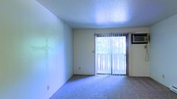 This single-floor space features a clean, straightforward layout centered around a well-lit main living area. The room is finished with wall-to-wall carpeting in a neutral gray-blue tone, providing a comfortable underfoot feel throughout. Walls are painted in a soft, light neutral hue, contributing to an open and airy atmosphere.

A prominent sliding glass door serves as the focal point of the main living area, fitted with vertical blinds that allow for adjustable light control. The door provides direct access to an outdoor space, with views of surrounding greenery visible through the glass, bringing a natural element into the interior. Natural light filters generously through the door, brightening the room throughout the day.

A wall-mounted air conditioning unit is positioned adjacent to the sliding door, offering a practical climate control solution. The ceiling features a textured popcorn finish, consistent with the walls' neutral palette. Electrical outlets are conveniently placed along the baseboards.

Overall, the space presents a functional and modest interior, well-suited for comfortable day-to-day living with its neutral finishes, natural light, and outdoor connectivity.