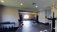The Spinning Room is a well-equipped, single-floor fitness space designed to support a variety of workout styles. The room is finished with neutral gray walls accented by a bold yellow architectural beam, lending the space a modern, energetic aesthetic. Durable rubber flooring covers the entire area, providing both safety and functionality for high-impact activities.

A rack of medicine balls in various weights lines one wall, accompanied by weight benches positioned toward the center of the room. Stationary cycling bikes are arranged in a dedicated section, making the space ideal for spin classes or individual cardio sessions. A standing punching bag adds a combat fitness element to the room's versatility.

Recessed lighting illuminates the space evenly throughout, while a large arched window at the far end introduces natural light into the interior. A set of white panel doors provides access to additional areas. The open layout allows for comfortable movement between equipment zones, and the overall design prioritizes function while maintaining a clean, motivating environment suited for group or individual training sessions.