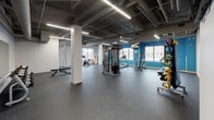 This fitness center offers a generously proportioned, open-plan workout space designed to accommodate a comprehensive range of exercise activities. The industrial-style aesthetic is defined by exposed concrete ceilings with visible ductwork, pipes, and linear LED lighting, lending the space a modern, urban character. Large windows along the perimeter walls invite natural light throughout, creating a bright and energizing atmosphere.

The main floor is covered in durable dark rubber flooring, practical for heavy equipment and high-intensity training alike. The space is organized to support a variety of fitness disciplines. Cardio equipment, including treadmills, elliptical machines, and a stationary spin bike, is distributed throughout the floor. A multi-station cable machine serves as a central strength training anchor, complemented by a rack of dumbbells, adjustable benches, and resistance training accessories.

A dedicated functional training area features balance and stability tools, resistance bands, and medicine balls on a rolling rack. Ceiling fans provide additional air circulation across the open floor. Interior windows connect to an adjacent office or reception area, supporting staff oversight. The overall layout is thoughtfully arranged to maximize usable floor space, making this fitness center a versatile and fully equipped facility suitable for a wide range of users and training styles.