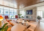 This single-floor lounge space is defined by a refined contemporary design that balances openness with functionality. The warm wood-paneled ceiling, fitted with recessed lighting, creates an inviting atmosphere while adding architectural character throughout the space. A wall of floor-to-ceiling windows along one side floods the interior with abundant natural light and provides expansive views of the surrounding cityscape, with sheer drapery offering adjustable privacy.

The main lounge area is anchored by a generously sized sectional sofa in neutral tones, accented with colorful throw pillows in warm reds, yellows, and corals. Organic-shaped white coffee tables and gold-accented table lamps add sculptural interest. A striking geometric area rug in deep burgundy, gold, and cream tones defines the seating zone and introduces a bold design element. Clusters of deep red high-back lounge chairs paired with round side tables provide additional intimate seating near the windows.

A stone-clad fireplace with a wall-mounted display screen anchors one end of the room. Glass-walled private rooms, accessible via sliding partitions, offer enclosed spaces for focused use. Light wood flooring transitions throughout, and lush greenery is positioned throughout to enhance the space's welcoming, sophisticated ambiance.