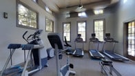 This fitness center offers a thoughtfully designed workout environment that balances functionality with a welcoming atmosphere. The space is bathed in natural light streaming through multiple large windows positioned along the walls, as well as higher clerestory windows near the ceiling, creating a bright and energizing setting for exercise. The neutral gray walls and carpeted flooring contribute to a clean, focused ambiance throughout. The cardio area is well-appointed with multiple treadmills, elliptical machines, and a stationary bike, all arranged to allow comfortable spacing between equipment. A ceiling fan is centrally positioned to promote air circulation during workouts. The elevated window placement ensures privacy while still allowing views of the surrounding greenery, connecting users to the outdoors without distraction. The overall layout is practical and efficient, making it easy to navigate between equipment. The single-floor design ensures accessibility, and the consistent finishes throughout the space maintain a cohesive, professional feel. This fitness center is well-suited for a variety of cardio-focused workout routines in a comfortable and naturally lit environment.