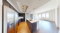 Located at 7911 Westpark Dr in McLean, Virginia, this well-appointed 2-bedroom, 2-bathroom residence offers a thoughtfully designed open floor plan. The combined kitchen and living area serves as the heart of the home, showcasing rich hardwood flooring, dark espresso cabinetry, and a large center island with a white quartz countertop and undermount sink, illuminated by modern pendant lighting. Stainless steel appliances, including a French door refrigerator, double oven range, and built-in microwave, complete the kitchen. A built-in desk and shelving unit add functional versatility to the space. The living area benefits from expansive floor-to-ceiling windows that flood the room with natural light and offer sweeping panoramic views of the surrounding McLean skyline. Both bedrooms feature plush carpeting and generous closet space. The primary bedroom connects directly to a full en-suite bathroom with dark wood cabinetry, a white dual-sink vanity, tile flooring, and a soaking tub with tiled surround. The second bathroom mirrors the refined finishes with matching cabinetry and a tub-shower combination. Throughout the residence, tall ceilings and a neutral color palette create a bright, airy ambiance.