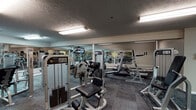 Located at 3500 Beaver Place Rd in Lexington, Kentucky, this fitness center offers a well-equipped, single-floor workout facility designed to support a full range of strength and conditioning exercises. The room features textured ceilings with recessed fluorescent lighting that evenly illuminates the entire space, creating a bright and functional environment throughout.

The facility is outfitted with a varied selection of Life Fitness selectorized strength machines, including cable systems, leg press, chest press, and multi-function tower units, catering to both upper and lower body training. Free weight benches and additional resistance equipment are distributed throughout the room, allowing for diverse workout routines. Cardio equipment, including treadmills, is also present and positioned along the mirrored walls.

Large wall-mounted mirrors span multiple sections of the fitness center, visually expanding the space and allowing users to monitor their form during exercise. The floor is covered in durable, patterned carpet tiles suited for a high-traffic gym environment. A staircase is visible in one corner of the room, indicating access to additional areas of the building. The neutral wall tones and structured layout contribute to a clean, organized, and practical atmosphere focused entirely on fitness.