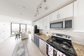 Located at 50-54 Sussex Avenue in Newark, NJ, this one-bedroom, one-bathroom unit offers a well-designed layout that maximizes every square foot of its compact footprint. The open galley-style kitchen is a standout feature, showcasing two-tone cabinetry with crisp white upper cabinets and slate-blue lower cabinets, granite countertops, a white subway tile backsplash, and a full suite of stainless steel appliances including a refrigerator, electric range, microwave, and dishwasher. Light wood-tone flooring runs throughout, lending warmth and continuity to the space. The kitchen flows naturally into the living area, which is anchored by a striking floor-to-ceiling window that floods the space with natural light and frames an open city view — a defining architectural feature of the unit. The bedroom is a cozy, efficiently sized retreat with darker wood-tone flooring, white walls, and a clean, minimal aesthetic. The bathroom is finished with large-format dark slate tile flooring, a white vanity with granite countertop, a framed mirror, and crisp white walls, creating a sleek, modern feel. Track lighting in the kitchen and an overall bright, neutral palette give the entire unit a fresh, contemporary character throughout.