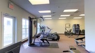 This fitness center offers a practical and well-organized space for exercise and wellness. The room is finished with a drop-panel ceiling fitted with recessed fluorescent lighting panels that provide bright, even illumination throughout. Neutral-toned walls and carpeted flooring in the main exercise area contribute to a clean, comfortable atmosphere, while a tiled section near the entrance provides a durable transition zone.

The space is equipped with an array of cardio machines, including elliptical trainers, treadmills, and a stationary recumbent bike, arranged to allow comfortable spacing between users. A multi-function cable strength training machine is positioned near the windows, making it a versatile option for resistance workouts. Additional equipment, including what appears to be a stair stepper, is situated toward the rear of the room.

Natural light enters through windows along one wall, adding a welcoming brightness to complement the overhead lighting. A framed piece of wall art and a small plant add subtle decorative touches that soften the functional nature of the space. The overall layout is efficient and user-friendly, with enough open floor space to accommodate stretching or floor exercises. This fitness center is well-suited for a range of fitness levels and exercise preferences.
