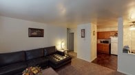 This single-floor unit offers a functional and straightforward layout with clearly defined living spaces. The living room features carpet flooring, a wall-mounted air conditioning unit, and a baseboard heating system, providing year-round climate control. Natural light enters through a large window fitted with horizontal blinds, creating a bright and comfortable atmosphere. A secondary area near the entry accommodates a media console with a wall-mounted television.

The kitchen is equipped with warm-toned oak cabinetry, a full-size refrigerator, microwave, and dark-tiled flooring, offering ample storage and counter space. Adjacent to the kitchen, the dining area is defined by a striking full-height exposed brick accent wall, adding a distinctive, textural element to the space. A ceiling light fixture illuminates the cozy dining nook, which includes a compact table and seating for two.

The unit includes two carpeted bedrooms, each featuring a window with blinds and baseboard heating. The primary bedroom offers generous closet space with built-in bifold doors. The second bedroom is well-suited as a nursery or additional sleeping quarters. The bathroom features tile flooring, a shower-tub combination, a vanity with storage, and recessed lighting.
