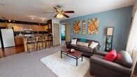 This single-floor residence offers a thoughtfully designed open layout that connects the living area and kitchen into a cohesive, comfortable living space. The living room features a bold teal accent wall, ceiling fan, soft carpeting, and a bright window that lets in natural light, creating a warm and inviting atmosphere. A second seating area provides additional space to relax. The two areas together offer generous room for entertaining and everyday living.

The kitchen is well-equipped with warm oak cabinetry, ample counter space, and hardwood flooring that adds a rich contrast to the dark-painted walls. Track lighting illuminates the workspace effectively, and the layout includes a full suite of appliances — refrigerator, stove, dishwasher, and microwave — along with a breakfast bar for casual dining.

The primary bedroom is spacious with plush blue carpet, a dedicated closet, and a convenient vanity area with a sink, offering a semi-private en-suite feel. A second bedroom features its own carpet and dedicated storage. Both bathrooms are finished with tile flooring and shower-tub combinations. The overall floor plan, visible in the dollhouse view, reveals a well-organized and efficiently arranged single-level home ideal for comfortable everyday living.