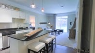 This single-floor space showcases an open-concept layout that seamlessly connects the kitchen and living areas. The kitchen features dark countertops on a peninsula-style island with seating for multiple guests on upholstered bar stools, making it an ideal gathering spot. Pendant lights with copper-toned fixtures hang above the island, adding a warm, industrial touch to the otherwise light and airy palette. White cabinetry lines the kitchen walls, complementing the soft blue-gray tones used throughout the space.

The living area flows naturally from the kitchen, offering a comfortable seating arrangement centered around a round coffee table with a woven base. A large sliding glass door floods the room with natural light and provides views of the exterior. Soft carpeting in the living zone contrasts with the dark hardwood-style flooring in the kitchen area, subtly defining each space. A floor lamp and tasteful décor accents contribute to the relaxed, welcoming ambiance. The overall design balances functionality with a clean, contemporary aesthetic throughout.