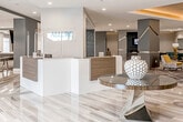 Allure is a thoughtfully designed, single-floor amenity space that exudes contemporary sophistication from the moment you enter. The reception area greets visitors with a sleek, two-toned desk finished in warm wood and crisp white, set against polished marble-look tile flooring that flows seamlessly throughout the space. A backlit "Allure" logo sign anchors the welcoming entry, establishing the refined aesthetic found throughout.

Beyond the reception, the space opens into a generously sized social lounge featuring coffered ceilings with warm ambient uplighting, a boldly patterned area rug, and an eclectic mix of navy, teal, and gold seating arrangements that invite gathering and relaxation. A dedicated bar-height counter runs along one wall, framed by a dark navy accent panel and a trio of flat-screen monitors — ideal for watching live events or presentations.

A large communal high-top table with textured swivel stools anchors a central flex area, bathed in natural light from oversized windows. Adjacent, a game room offers a full-size billiards table set atop high-gloss tile flooring beneath recessed lighting.

A private meeting room provides an intimate setting with a round black table, upholstered chairs, and vertically slit mirror accents along textured walls, creating a refined atmosphere for focused gatherings. The overall design balances bold, modern aesthetics with functional, community-centered spaces.