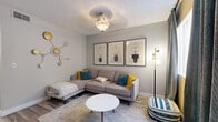 This two-story, 2-bedroom, 2-bathroom residence in Chandler, Arizona offers a thoughtfully designed layout spread across two floors. The main level features a welcoming living room with wood-look flooring, a textured ceiling, and a decorative pendant light fixture. The space is accented by eclectic wall art and large draping curtains that frame a generous window, filling the room with natural light. The kitchen is a standout, featuring two-tone cabinetry — crisp white uppers paired with gray lowers — complemented by white quartz countertops, stainless steel appliances, and a deep undermount sink. Wood-look flooring carries throughout the main level, tying the spaces together cohesively. Both bathrooms are finished with gray vanity cabinetry, quartz countertops, and wood-look flooring; one bathroom includes a soaking tub with a decorative curtain. The two bedrooms are carpeted and bathed in natural light from large windows with vertical blinds. The primary bedroom offers a comfortable, well-proportioned retreat, while the second bedroom mirrors the same calm, neutral aesthetic. A staircase connects the two levels, and a hallway and closet provide practical storage. The overall ambiance is modern, clean, and inviting throughout.