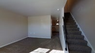 This two-story residence offers a functional and straightforward layout designed for comfortable everyday living. The main level features a spacious living area finished with wall-to-wall carpeting that adds warmth underfoot. Neutral-toned walls keep the space bright and versatile, while natural light filters in through a front-facing window near the entryway, casting a natural glow across the room. An open sightline connects the living area to the kitchen, which is visible in the background and features warm wood-toned cabinetry, providing a welcoming sense of continuity between spaces. A dedicated entryway with a half-lite door allows natural light to enter the front of the home. A carpeted staircase with a sleek black metal railing rises along one side of the main level, leading to the second floor and adding a defined vertical element to the interior. The overall ambiance is clean, neutral, and move-in ready, with a simple design aesthetic that allows residents to personalize the space with ease. The two-floor layout provides a natural separation between living and private areas, making this a well-structured home suited to a variety of lifestyles.