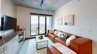 Located at 708 S Sixth St in Champaign, Illinois, this contemporary multi-bedroom unit showcases a thoughtfully designed interior that blends industrial and modern aesthetics throughout. Exposed concrete ceilings and structural columns lend a distinctive urban character to the space, while light wood flooring in the main living areas adds warmth and cohesion.

The open-concept kitchen is outfitted with stainless steel appliances — including a range, microwave, dishwasher, and full-size refrigerator — complemented by two-tone cabinetry featuring white upper cabinets and wood-finish lower cabinets, dark countertops, and a striking penny-tile backsplash. A kitchen island with a butcher-block top provides additional prep and dining space.

The living area is bright and airy, anchored by large floor-to-ceiling windows that frame sweeping elevated views of the Champaign cityscape. Ceiling-mounted track lighting enhances the industrial feel of the space.

Each bedroom is furnished with a bed and a built-in or freestanding wood desk, making them well-suited for both rest and study. Select bedrooms feature windows with impressive elevated city views and ceiling fans. The bathrooms are finished with wood-tone vanities, undermount sinks, and modern fixtures; one includes a glass-enclosed walk-in shower with tiled walls.