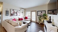 This efficiently designed single-floor residence offers 1 bedroom and 2 bathrooms within a smart, open-concept layout. The living room features rich hardwood-style flooring, a wall-mounted mini-split HVAC unit, large windows with blinds that allow abundant natural light, and a glass door providing outdoor access. The space is complemented by contemporary wall art and a sectional seating arrangement that maximizes comfort.

The kitchen is well-equipped with warm walnut-toned cabinetry, stainless steel appliances including a refrigerator with ice maker, electric range, and built-in microwave. A broad white countertop peninsula serves as both a prep surface and casual dining space, with hardwood-style flooring continuing throughout.

The dining area accommodates a glass-top round table with upholstered seating beneath a classic chandelier, enhanced by world-map wall art that adds a cosmopolitan character to the space.

The bedroom is a cozy, carpeted retreat with a window offering natural light, providing a quiet atmosphere for rest.

Both bathrooms are finished with warm wood vanities, white countertops, wide mirrors with bar lighting, and hardwood-style flooring, offering clean and functional designs. A dedicated closet completes this well-organized floor plan.