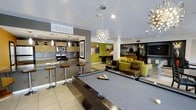 This single-floor community lounge space is designed with a modern aesthetic that blends functionality and style seamlessly. The open layout integrates several distinct zones, creating a versatile environment ideal for socializing and recreation. At the heart of the space is a full-size billiards table, positioned beneath a striking starburst chandelier that serves as a focal point and adds a bold design statement to the room. Adjacent to the billiards area is a well-appointed kitchenette, featuring dark countertops, warm wood cabinetry, a tile backsplash, and stainless steel appliances including a microwave and range. A kitchen bar with modern swivel stools provides casual seating and a natural gathering spot. Pendant lighting above the bar complements the overall lighting scheme. The lounge area is furnished with contemporary chartreuse and chocolate-toned sofas and seating, accented by vibrant green walls and colorful artwork that energize the space. A built-in shelving unit in the background adds both display and storage functionality. The light tile flooring and recessed lighting throughout contribute to a bright, open ambiance that ties all areas of the space together cohesively.