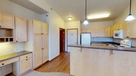 This single-floor space showcases an open-concept kitchen and living area defined by light maple wood cabinetry throughout. The kitchen is well-appointed with a peninsula-style breakfast bar featuring a dark laminate countertop, offering both additional prep space and a casual seating option. A pendant light hangs above the bar, complementing the flush-mount ceiling fixtures that illuminate the space. Full-height pantry-style cabinets provide generous storage along one wall, while upper and lower cabinets line the kitchen perimeter, maximizing organization. A white refrigerator anchors one end of the kitchen, and a sink is positioned along the far wall. The vaulted ceiling adds a sense of openness and volume, drawing the eye upward and enhancing the airy feel of the interior. Warm-toned hardwood-style flooring runs throughout the main area, transitioning to carpet in adjacent spaces. Neutral wall tones keep the interior feeling clean and versatile. A hallway visible from the kitchen leads to additional rooms, and a six-panel interior door adds a classic architectural detail. Overall, the space presents a practical and cohesive design suited for everyday living.