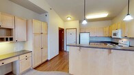 This single-floor space showcases an open-concept kitchen and living area defined by light maple wood cabinetry throughout. The kitchen is well-appointed with a peninsula-style breakfast bar featuring a dark laminate countertop, offering both additional prep space and a casual seating option. A pendant light hangs above the bar, complementing the flush-mount ceiling fixtures that illuminate the space. Full-height pantry-style cabinets provide generous storage along one wall, while upper and lower cabinets line the kitchen perimeter, maximizing organization. A white refrigerator anchors one end of the kitchen, and a sink is positioned along the far wall. The vaulted ceiling adds a sense of openness and volume, drawing the eye upward and enhancing the airy feel of the interior. Warm-toned hardwood-style flooring runs throughout the main area, transitioning to carpet in adjacent spaces. Neutral wall tones keep the interior feeling clean and versatile. A hallway visible from the kitchen leads to additional rooms, and a six-panel interior door adds a classic architectural detail. Overall, the space presents a practical and cohesive design suited for everyday living.
