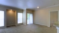 This single-story space features a well-proportioned living area finished with wall-to-wall carpeting in a neutral tone, creating a comfortable and cohesive feel underfoot. Crown molding lines the perimeter of the ceiling, adding a refined architectural detail that elevates the overall aesthetic. The walls are painted in a warm beige, contributing to a bright and inviting ambiance throughout.

Natural light is a standout feature of this room, delivered through two sources: a large fixed window along one wall and a full-height sliding glass door fitted with vertical blinds. The sliding door provides direct access to an outdoor area, where glimpses of mature greenery can be seen, enhancing the sense of connection to the outside. A recessed light fixture in the ceiling provides supplemental illumination when needed.

The layout is open and unobstructed, offering flexibility for a variety of furniture arrangements. White baseboards frame the room cleanly, complementing the crown molding above. A small niche or recessed section is visible near the window wall, adding a subtle architectural interest. Overall, the space presents a clean, move-in ready environment with classic detailing and ample natural light.