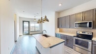 This thoughtfully designed one-bedroom, one-bathroom residence offers a modern open-concept layout that maximizes its well-utilized floor area. The kitchen serves as the centerpiece of the living space, showcasing a large granite-topped island with an undermount sink, dramatic pendant lighting in a brushed brass finish, and custom gray shaker-style cabinetry. A mosaic tile backsplash, stainless steel appliances — including a range, microwave, and refrigerator — and a built-in wine cooler complete the kitchen's refined character. Light wood-look flooring extends throughout the main living area, enhancing the bright, airy ambiance. Large windows frame elevated city views, filling the space with abundant natural light. The bedroom is carpeted for comfort and features a ceiling fan, a window with skyline views, and access to a well-organized walk-in closet outfitted with multi-tiered hanging rods and built-in shelving. The bathroom is finished with wood-plank porcelain tile flooring, gray vanity cabinetry with granite countertops, a soaking tub, and a separate tiled shower. A hallway with a teal accent door and a dedicated closet provide additional practicality and character to this polished urban residence.