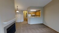 This single-floor residence showcases an open-concept design that seamlessly connects the living area and kitchen. The living room is anchored by a brick-framed fireplace, adding a warm focal point to the space, and is finished with wall-to-wall carpet in neutral tones that extends throughout the unit. A pendant light fixture adds a decorative touch near what appears to be a closet or storage area with bifold doors.

The kitchen is defined by a raised peninsula that separates it from the living space, offering a natural divider while maintaining an open feel. Light oak cabinetry lines the upper walls, providing ample storage, while white appliances—including a refrigerator and range—equip the space for everyday cooking. Fluorescent overhead lighting illuminates the kitchen workspace efficiently.

A hallway visible from the main living area leads toward additional rooms, suggesting a well-organized floor plan. The overall color palette consists of soft beige and warm gray tones, creating a cohesive and understated aesthetic throughout. The layout prioritizes functional flow, making the space feel connected and accessible.