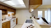 This two-story residence offers a practical and well-organized layout across two levels connected by a carpeted staircase fitted with a wooden handrail. The stairway is positioned adjacent to the main living area, providing easy access between floors while maintaining an open sightline into the kitchen below.

The kitchen is thoughtfully designed for functionality, featuring warm wood cabinetry that provides ample storage space. White appliances, including a range, complement the light countertops that run along the walls, offering generous prep space. A double stainless steel sink is centrally positioned along the counter, and a built-in overhead light panel brightens the workspace from above. A pass-through window or counter opening connects the kitchen to the adjacent living area, enhancing the flow between spaces. A door with a glass pane at the end of the kitchen allows natural light to filter in.

The living areas are finished with light-toned carpet that adds warmth and comfort underfoot. The overall interior palette is kept neutral with white walls throughout, creating a clean, bright atmosphere. The multi-level layout provides a clear separation between the main living spaces on the lower floor and the upper level, making this a well-structured and versatile home.
