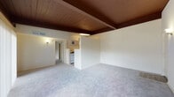 This single-floor residence showcases a distinctive mid-century modern aesthetic, anchored by rich, dark wood-planked ceilings with exposed beams that run throughout the main living areas and bedroom, lending warmth and architectural character to the space. The generous living room is finished with plush light-gray carpeting and features a sliding glass door that opens to a private balcony, inviting natural light and outdoor views into the interior. A half-wall partition separates the living area from the kitchen zone, maintaining an open feel while defining each space. The galley-style kitchen is well-appointed with warm honey-toned wood cabinetry, dark granite-style countertops, a stainless sink, and white appliances including a refrigerator and range. Light wood-look flooring in the kitchen provides a clean contrast to the surrounding carpeted areas. The bedroom continues the signature beamed wood ceiling and offers generous wall space, plush carpeting, a spacious reach-in closet, and direct access to the bathroom. The bathroom is clean and functional, featuring a full soaking tub with tiled surround, a white vanity with under-cabinet storage, a large mirror, and coordinating light wood-look flooring. Overall, the space presents a cohesive, retro-inspired design with a bright, neutral palette throughout.
