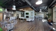 This expansive single-floor amenity lounge showcases a striking blend of industrial and contemporary design. The soaring dark-painted ceiling features exposed ductwork and mechanical systems, lending the space an urban, loft-inspired character. Statement pendant lights hang at varying heights, casting a warm, inviting glow across the rich hardwood flooring that runs throughout.

At the heart of the space is a central feature wall housing a sleek fireplace topped by a wall-mounted flat-screen television, framed by curated wall art. Surrounding this focal point is a generously sized seating area outfitted with modern white sofas, accent chairs, and coffee tables — ideal for casual gatherings or quiet lounging. A warm wood slat ceiling detail above this zone adds a refined contrast to the industrial aesthetic overhead.

Toward one end of the space, a bar-height counter with white stools provides a dedicated spot for socializing, complemented by what appears to be a café or beverage station. On the opposite side, a billiards table anchors a game area, adding a recreational dimension to the lounge. Large windows along the perimeter draw in abundant natural light, contributing to the open, airy atmosphere of this thoughtfully designed communal space.