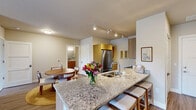 This thoughtfully designed single-floor residence offers a cohesive layout that balances functionality and comfort throughout. The heart of the home is an open-concept kitchen and dining area, where warm wood cabinetry pairs beautifully with granite countertops and a large breakfast bar with seating. Stainless steel appliances, a double-basin sink, and track lighting complete the kitchen's polished, modern aesthetic. A round dining table nearby provides an intimate space for meals.

The living room is bright and inviting, featuring wood-look flooring, track lighting, and a large sliding glass door that opens to a private balcony with outdoor seating — bringing in natural light and views of the surrounding greenery. A wall-mounted flat-screen TV and a rustic media console add to the room's relaxed, stylish character.

The bedroom offers a serene retreat with neutral tones, warm layered bedding, and soft ambient lighting. Adjacent is a dedicated home office, bathed in natural light from a large window, and configured for productive work-from-home use with a substantial desk and shelving unit.

The bathroom features a clean, contemporary design with a walk-in glass-panel shower, built-in niche shelving, and neutral tile flooring, creating a calm and functional space. Light-toned walls and white trim carry a consistent, airy palette throughout the entire residence.