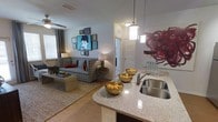 This single-floor residence showcases a thoughtfully designed open-concept layout that integrates the kitchen and living area into one cohesive, inviting space. The kitchen features sleek granite countertops with a double stainless steel sink, pendant lighting that adds a contemporary touch, and a curved breakfast bar that serves as a natural transition into the living space. Decorative elements, including bold red sculptural accents and coordinated bowl arrangements, lend a curated aesthetic to the kitchen area. The living room is warm and welcoming, anchored by hardwood-style flooring and a large area rug that defines the seating zone. A patterned sofa, layered wall art — highlighted by a striking pop-art portrait — and a ceiling fan overhead contribute to the room's eclectic yet comfortable character. Natural light filters in through window treatments, while ambient lamps provide a soft, cozy glow. A hallway leading to additional rooms is visible in the background, hinting at the full extent of the floor plan. Overall, the space balances modern finishes with personal touches, creating a livable and visually engaging environment.