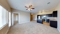 This single-floor space showcases an open-concept design that seamlessly connects the living area to the kitchen. The living space is finished with plush, light-toned carpeting and painted in neutral beige tones, creating a warm and inviting atmosphere. A ceiling fan with an integrated light fixture is centrally mounted, providing both air circulation and ambient lighting throughout the main living area.

The kitchen is positioned to the right and features dark espresso-toned cabinetry paired with stainless steel appliances, including a refrigerator, electric range, and overhead microwave. A kitchen island with a dark countertop adds additional prep and storage space. Track lighting above the kitchen ensures well-lit task areas. The kitchen floor transitions to hardwood or wood-look flooring, providing a practical and visually distinct contrast to the carpeted living space.

A hallway visible toward the back of the unit leads to additional rooms, suggesting a functional layout with clearly defined living and private zones. White trim and baseboards throughout the space add a clean, finished look.